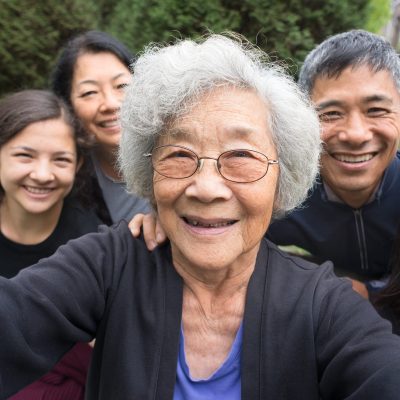 A fit ,senior, Asian Grandmother smiles with her children and mixed-ethnic grandchildren while posing for a selfie in a park garden.   Real, three generation family including a senior woman, mature adult son and daughter and teenage grand-daughters.  An assisted living facility can be seen in the background.  Camera point of view.